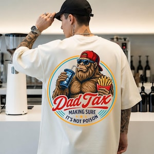 May include: An off-white short-sleeve t-shirt featuring a graphic on the back of a brown sasquatch wearing a red cap and blue sunglasses, holding a blue drink and red carton of fries, with the text "Dad Tax MAKING SURE IT'S NOT POISON".