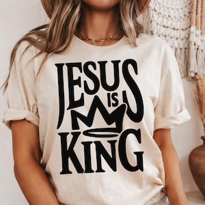 May include: A cream-coloured t-shirt featuring the text "JESUS IS KING" in bold black lettering, with a crown graphic. The shirt has short sleeves and a relaxed fit.