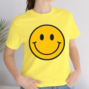 May include: Yellow t-shirt featuring a large, smiling emoji with a black outline. The emoji has two black eyes and a curved black mouth. The shirt is a solid yellow color.