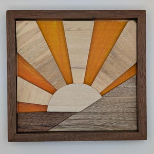 Puede incluir: Arte mural de madera con un diseño de amanecer. La obra de arte está compuesta por varios tonos de madera, incluyendo beige claro, naranja y marrón oscuro, dispuestos en un patrón geométrico. Enmarcado en un marco de madera marrón oscuro.
