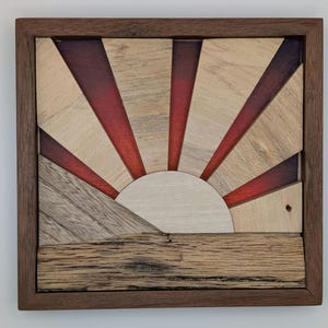 Puede incluir: Arte mural de madera con un diseño de amanecer. La obra de arte está enmarcada en madera marrón oscuro y muestra un sol con rayos en tonos rojos, beige y grises. El sol es de color beige claro.