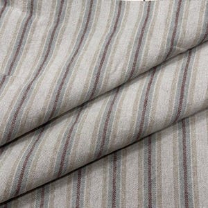 May include: Close-up of a folded fabric with vertical stripes. The stripes alternate between light beige, muted red, and light blue-gray. The fabric has a slightly textured appearance, suggesting a natural fiber like linen or cotton.