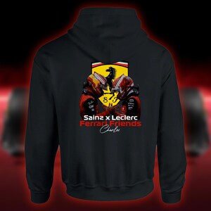Może przedstawiać: Czarna bluza z kapturem z grafiką dwóch kierowców wyścigowych w czerwonych i żółtych kaskach z logo Ferrari. Tekst brzmi "Sainz x Leclerc Ferrari Friends" i "Charles".