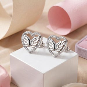 May include: A pair of silver-coloured heart-shaped stud earrings. Each earring features a wing design in the centre, surrounded by a halo of small, clear crystals. The earrings are displayed on a white surface against a soft-focus background.