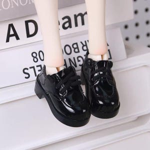 Puede incluir: Un par de zapatos de muñeca negros brillantes con cordones. Los zapatos tienen un tacón bajo y una punta redondeada. Los zapatos están sobre una superficie blanca. La palabra "AD" es visible en un libro en el fondo.