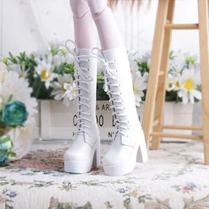 May include: White, knee-high boots with thick platform soles and block heels. The boots feature a lace-up design with white laces. The image shows the boots on a floral patterned surface, with a wooden structure and flowers in the background.