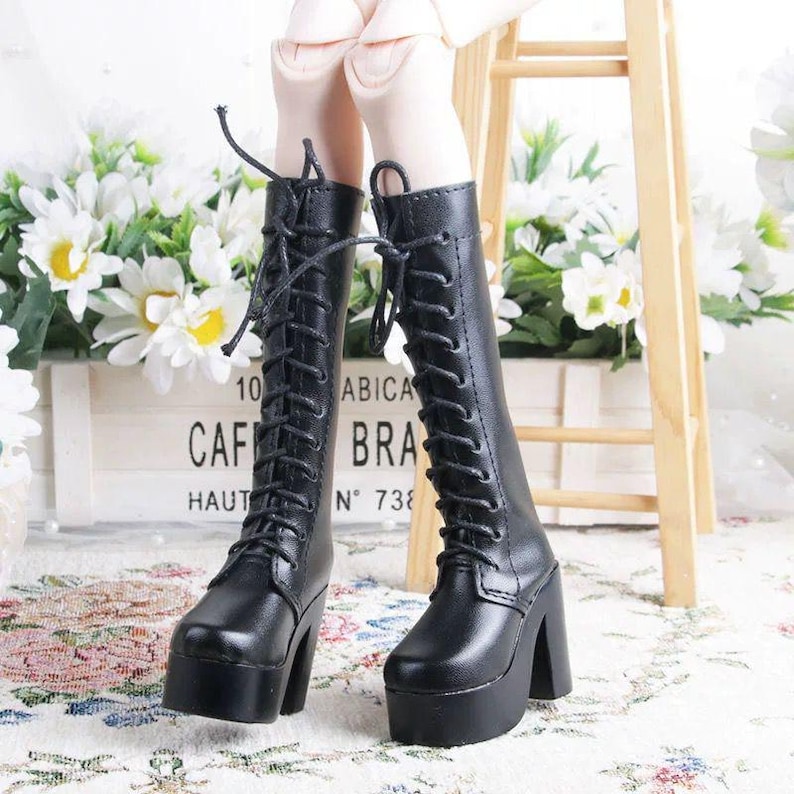 May include: Black, lace-up platform boots with thick heels. The boots have a rounded toe and are made of a smooth material. The boots are part of a doll's outfit. The background includes white flowers and a wooden sign.