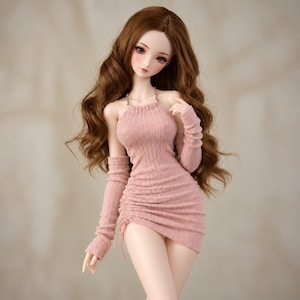Può includere: Una bambola snodabile con lunghi capelli castani ondulati, che indossa un miniabito a coste color rosa polvere con scollo all'americana e scaldabraccia lunghi abbinati. L'abito presenta un dettaglio arricciato laterale con coulisse e catenina dorata.