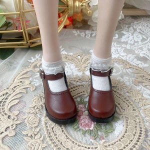 Pu&ograve; includere: Un paio di scarpe Mary Jane in pelle marrone con chiusura a fibbia, indossate con calzini bianchi rifiniti in pizzo. Le scarpe hanno la punta arrotondata e un piccolo tacco. Le scarpe sono su una superficie decorativa.