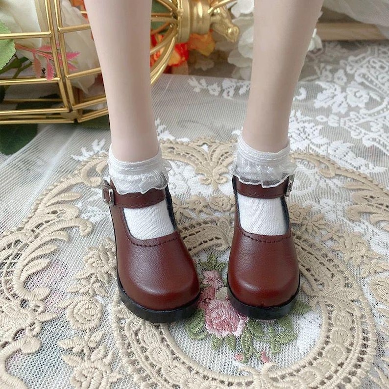 Pu&ograve; includere: Un paio di scarpe Mary Jane in pelle marrone con chiusura a fibbia, indossate con calzini bianchi rifiniti in pizzo. Le scarpe hanno la punta arrotondata e un piccolo tacco. Le scarpe sono su una superficie decorativa.