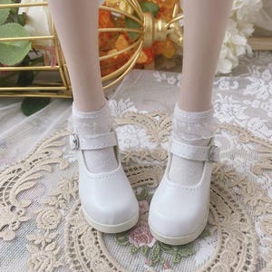 Pu&ograve; includere: Un paio di scarpe bianche stile Mary Jane con chiusura a fibbia. Le scarpe sono abbinate a calzini bianchi con bordi in pizzo. Le scarpe sono su una superficie a motivi floreali.