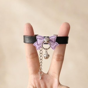 May include: Black leather choker featuring a lavender bow, silver bell, and chain. The bow has a scalloped edge. The bell and chain are silver. The choker is held up by fingers.