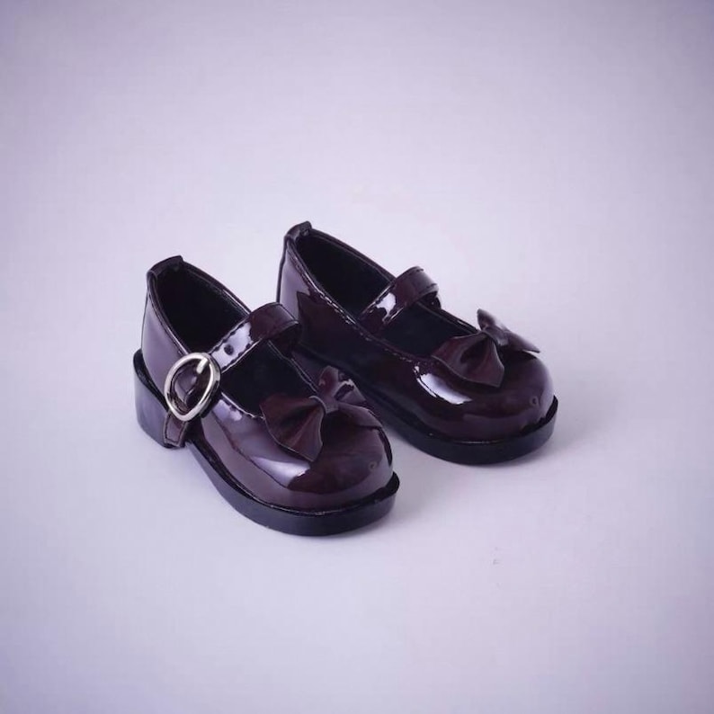 May include: A pair of dark burgundy patent leather Mary Jane shoes featuring a glossy finish, a rounded toe, a decorative bow on the front, and a single adjustable strap across the top secured with a silver-toned metal buckle and eyelet.