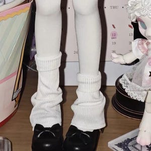 May include: A pair of white ribbed leg warmers worn over white tights, paired with black rounded-toe shoes featuring a small bow detail on the front. The leg warmers are scrunched at the ankles and extend up the lower legs.