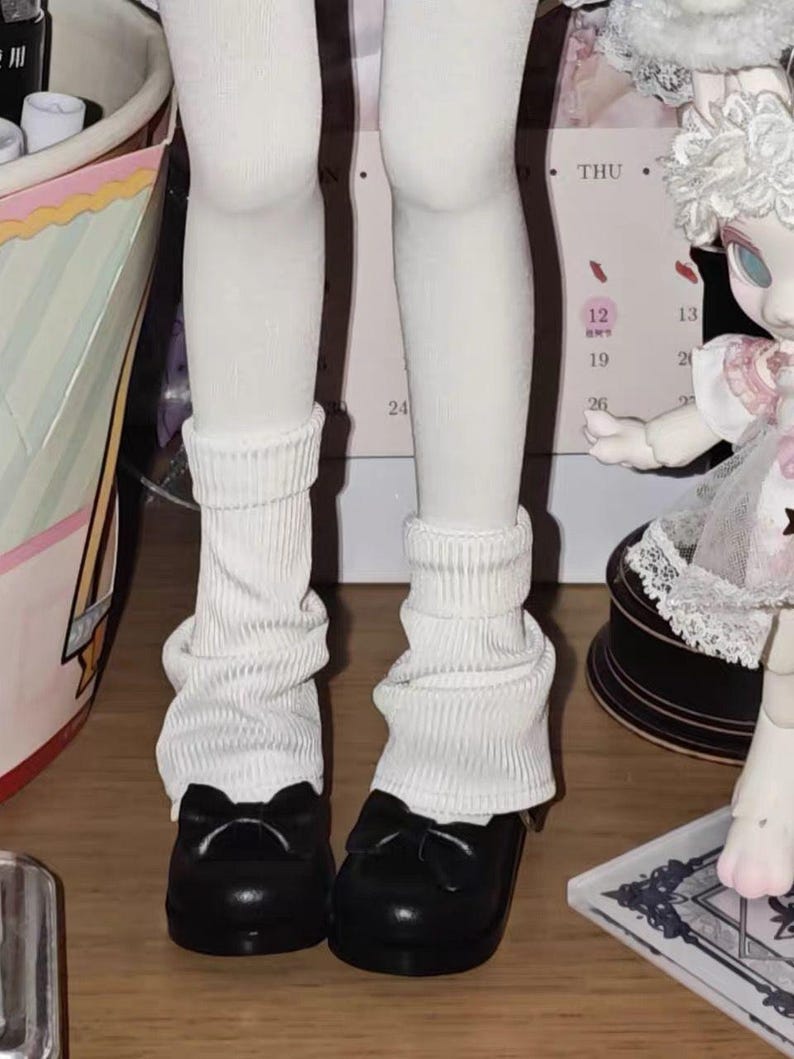 May include: A pair of white ribbed leg warmers worn over white tights, paired with black rounded-toe shoes featuring a small bow detail on the front. The leg warmers are scrunched at the ankles and extend up the lower legs.