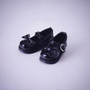 May include: A pair of black patent leather Mary Jane shoes featuring a rounded toe, a decorative bow on the front, and a single strap across the instep secured with a silver-toned metal buckle. The shoes have a smooth, glossy finish throughout.
