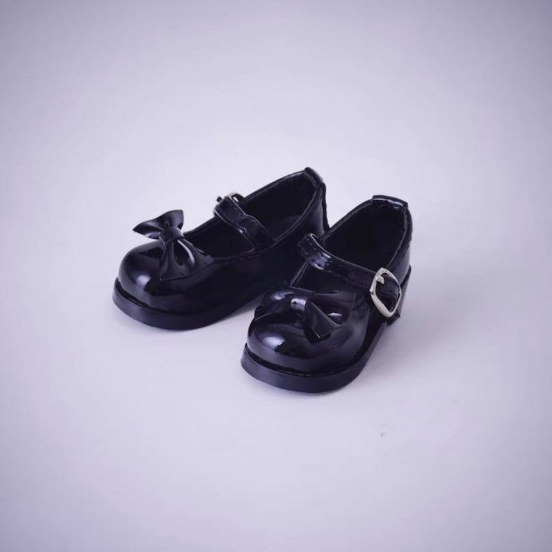 May include: A pair of black patent leather Mary Jane shoes featuring a rounded toe, a decorative bow on the front, and a single strap across the instep secured with a silver-toned metal buckle. The shoes have a smooth, glossy finish throughout.