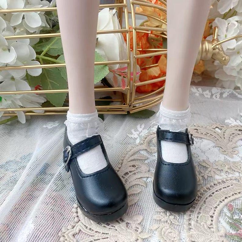 Pu&ograve; includere: Un paio di scarpe Mary Jane nere con cinturino singolo e fibbia color argento, indossate con calzini bianchi con volant. Le scarpe presentano una punta arrotondata e una finitura liscia, adatte per una bambola.