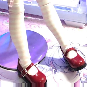 May include: A pair of glossy, dark red Mary Jane shoes with a buckle closure. The shoes are worn with striped white and pink socks. The shoes have a small heel and a rounded toe. The shoes are on a purple surface.