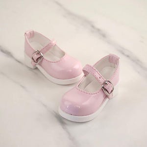 May include: A pair of light pink Mary Jane shoes with a glossy finish, featuring a rounded toe, a single strap across the instep with a silver-toned metal buckle, and a solid white sole. The interior lining of the shoes is white.