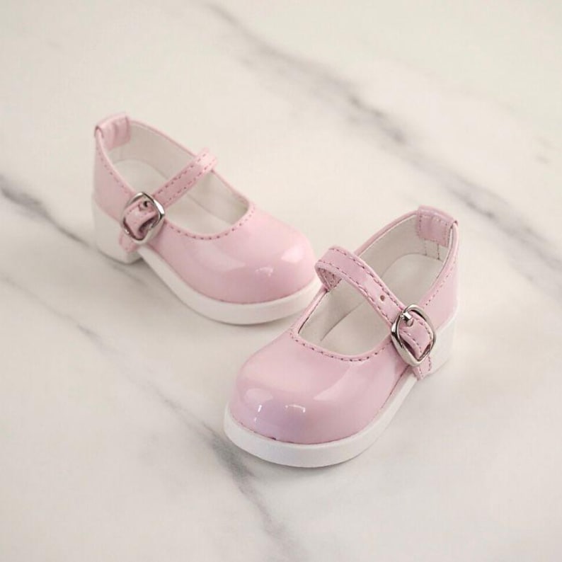 May include: A pair of light pink Mary Jane shoes with a glossy finish, featuring a rounded toe, a single strap across the instep with a silver-toned metal buckle, and a solid white sole. The interior lining of the shoes is white.