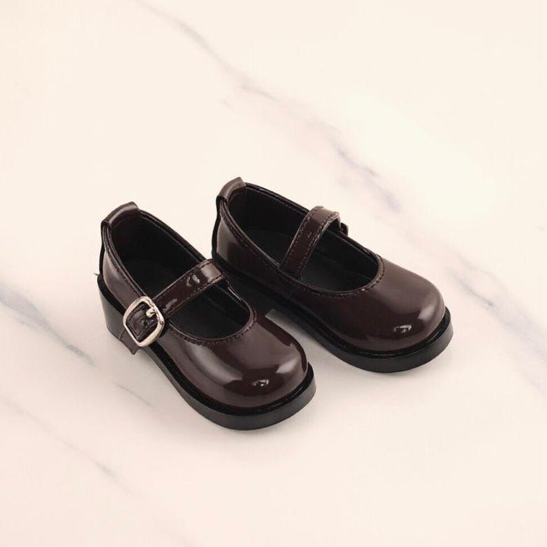 May include: A pair of glossy, dark brown Mary Jane style shoes. Each shoe features a strap with a silver buckle and a rounded toe. The shoes have a low, block heel and a thick sole, suitable for everyday wear.