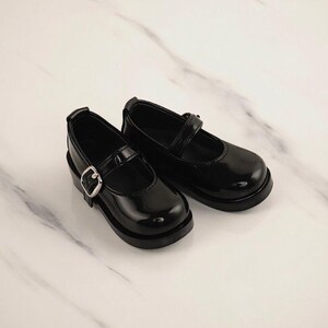 May include: A pair of black patent leather Mary Jane shoes with a silver buckle closure. The shoes have a rounded toe and a thick sole. The shoes are designed for children.