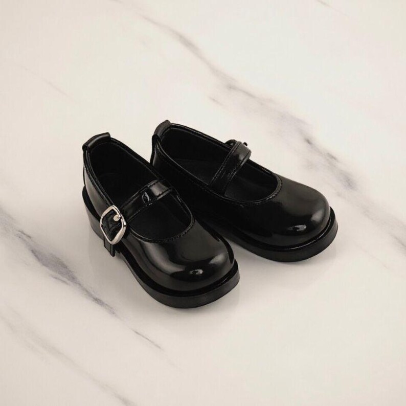 May include: A pair of black patent leather Mary Jane shoes with a silver buckle closure. The shoes have a rounded toe and a thick sole. The shoes are designed for children.