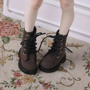 Puede incluir: Un par de botas marrones en miniatura con cordones y ojales negros. Las botas tienen un exterior marrón oscuro y una suela negra. Estas botas están diseñadas para una muñeca o una figura pequeña similar.
