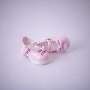 May include: A pair of light pink doll shoes featuring a rounded toe, a single strap across the instep with a small silver-toned metal buckle, and a decorative bow detail on the front. The shoes have a smooth finish and a white interior lining.