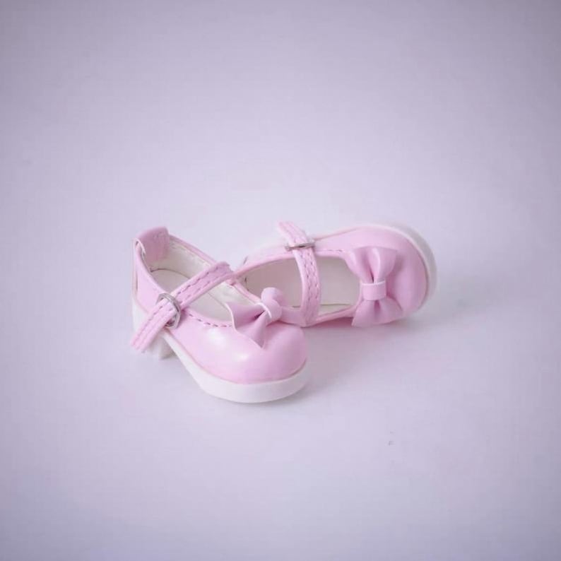 May include: A pair of light pink doll shoes featuring a rounded toe, a single strap across the instep with a small silver-toned metal buckle, and a decorative bow detail on the front. The shoes have a smooth finish and a white interior lining.