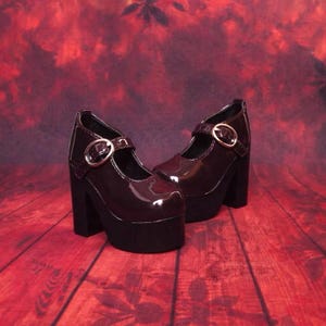 Puede incluir: Un par de zapatos Mary Jane de color burdeos oscuro con tacón de bloque alto y suela de plataforma gruesa. Cada zapato presenta un acabado de charol brillante, punta redondeada y una correa ajustable en el empeine con hebilla metálica dorada.