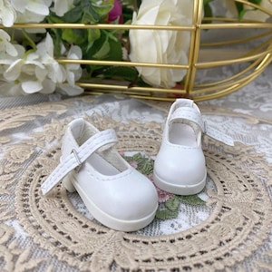 Pu&ograve; includere: Un paio di scarpe bianche da bambola in stile Mary Jane. Ogni scarpa ha un cinturino con fibbia argentata e punta arrotondata. Le scarpe sono realizzate in materiale liscio e opaco e poggiano su una suola bianca.
