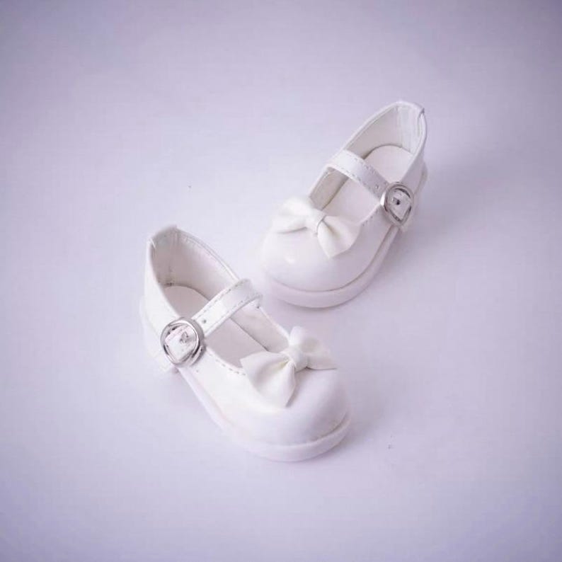 May include: A pair of white Mary Jane shoes featuring a smooth finish, a rounded toe, and a decorative bow on the front strap. Each shoe includes an adjustable strap with a silver-toned metal buckle closure, providing a secure fit and a clean look.