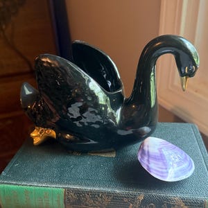 May include: A glossy black ceramic swan figurine with gold-painted beak and feet details, positioned on a dark green hardcover book next to a small, smooth, purple and white seashell. The swan has an open cavity on its back for holding items.