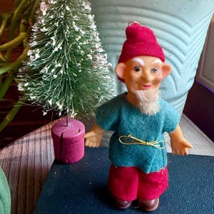 May include: A vintage gnome figurine with a red hat, teal shirt, and red pants. The gnome has a white beard and is standing next to a small, flocked Christmas tree. A gold-colored wire belt is around the gnome's waist. The background includes a large, light blue planter with green plants.