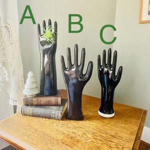 May include: Three black ceramic hand-shaped ring holders. The middle holder is the largest and the left holder has a small green plant inside.