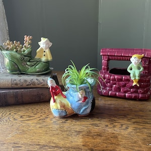 May include: Three ceramic planter pots shaped like shoes, each with a small gnome figurine. The green shoe has a plant growing out of it, the blue shoe has a small air plant, and the red shoe has a small gnome sitting on the side.