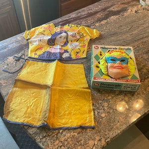 May include: A vintage fairy princess costume set for children. The set includes a yellow skirt, a top with a printed image of a fairy princess, and a plastic mask. The mask is blue, green, and yellow with a crown and a masquerade mask design. The box reads "Masquerade Costume".