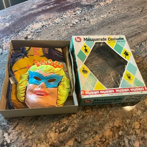 May include: A vintage Halloween costume box set with a plastic mask of a woman wearing a blue eye mask and a crown. The box is labeled "Masquerade Costume" with a black cat and witch silhouette. The text "Kusan Kusan Kusan Kusan" is printed on the box.