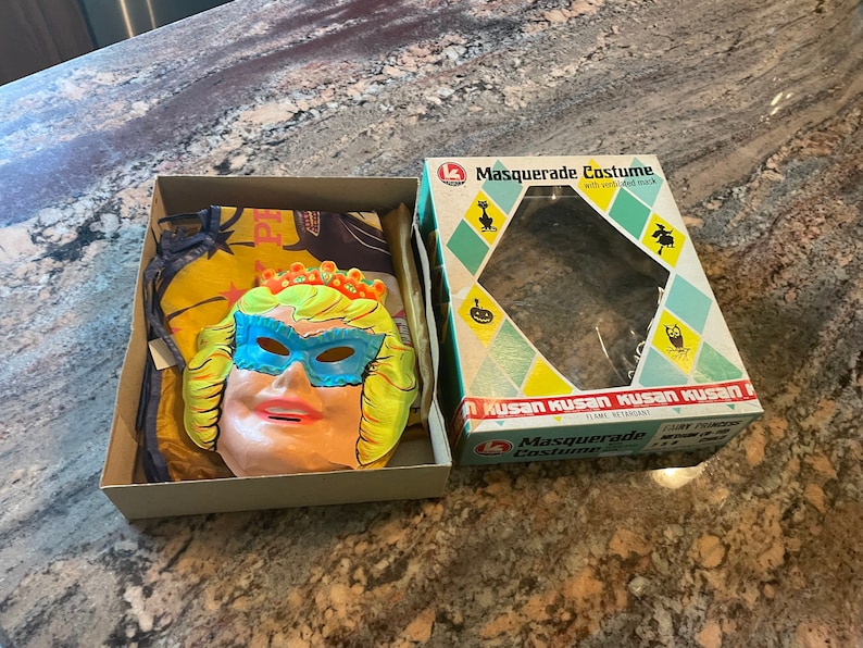 May include: A vintage Halloween costume box set with a plastic mask of a woman wearing a blue eye mask and a crown. The box is labeled "Masquerade Costume" with a black cat and witch silhouette. The text "Kusan Kusan Kusan Kusan" is printed on the box.