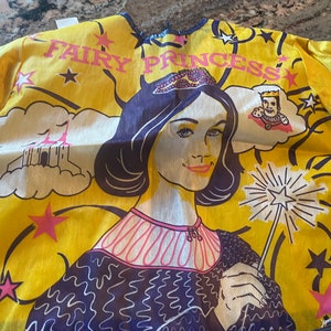 May include: A yellow fabric costume with a fairy princess design. The costume features a woman with a crown, a castle, and stars. The text "Fairy Princess" is printed on the costume.