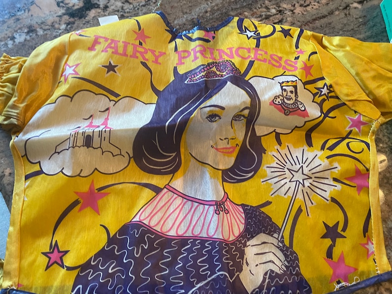 May include: A yellow fabric costume with a fairy princess design. The costume features a woman with a crown, a castle, and stars. The text "Fairy Princess" is printed on the costume.
