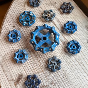 May include: A collection of vintage blue and gray valve handles arranged on a light-colored wooden surface. The handles vary in size and design, showcasing a rustic, industrial aesthetic. The handles are made of metal and have a weathered appearance.