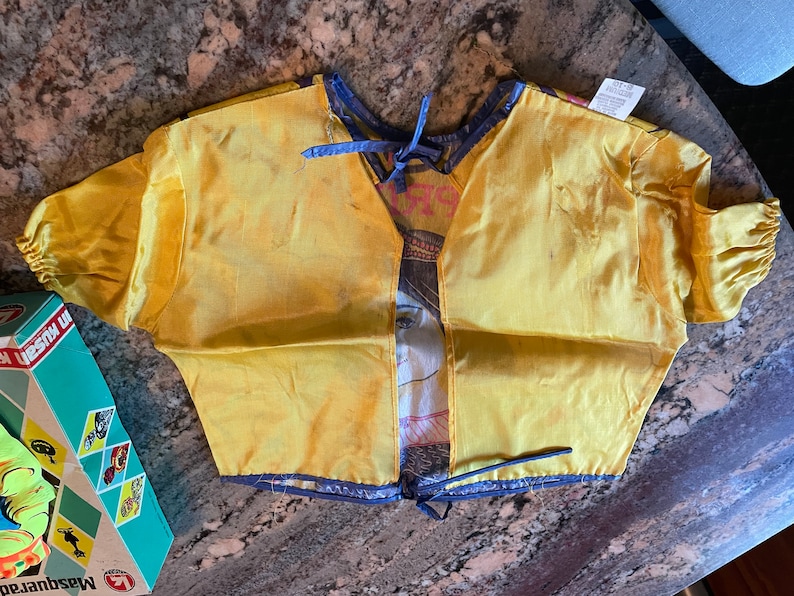 May include: A yellow satin costume top with a blue trim and a tie at the neck. The top has a printed design of a woman's face and a floral pattern.