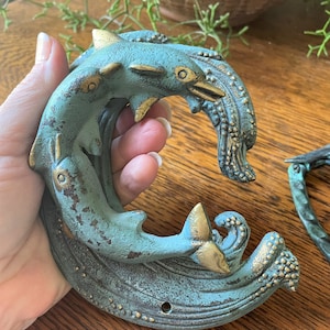May include: A teal and gold dolphin door knocker with a textured finish. The dolphins are intertwined and appear to be swimming in a wave.