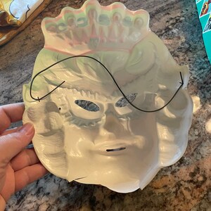 May include: White plastic masquerade mask with a black elastic band. The mask has a crown design on the top and eye holes with a black plastic eye mask.