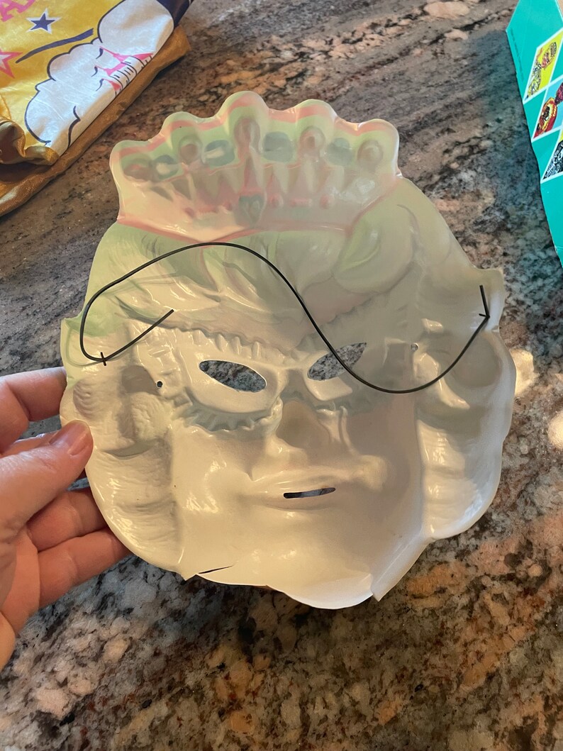 May include: White plastic masquerade mask with a black elastic band. The mask has a crown design on the top and eye holes with a black plastic eye mask.