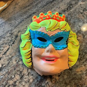 May include: A vintage plastic Halloween mask of a woman wearing a blue eye mask and a yellow crown. The mask has a pink mouth and a light pink face.