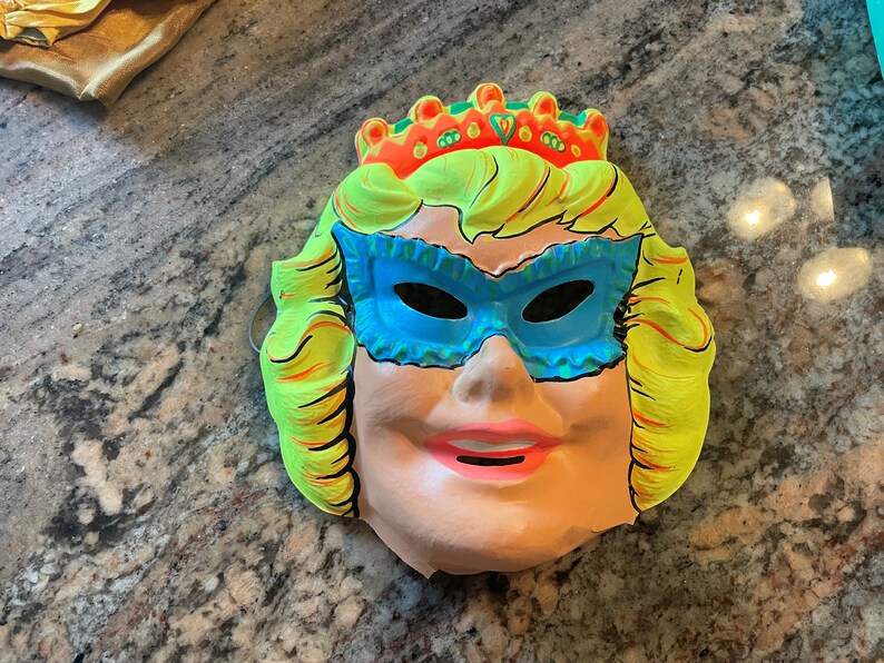 May include: A vintage plastic Halloween mask of a woman wearing a blue eye mask and a yellow crown. The mask has a pink mouth and a light pink face.
